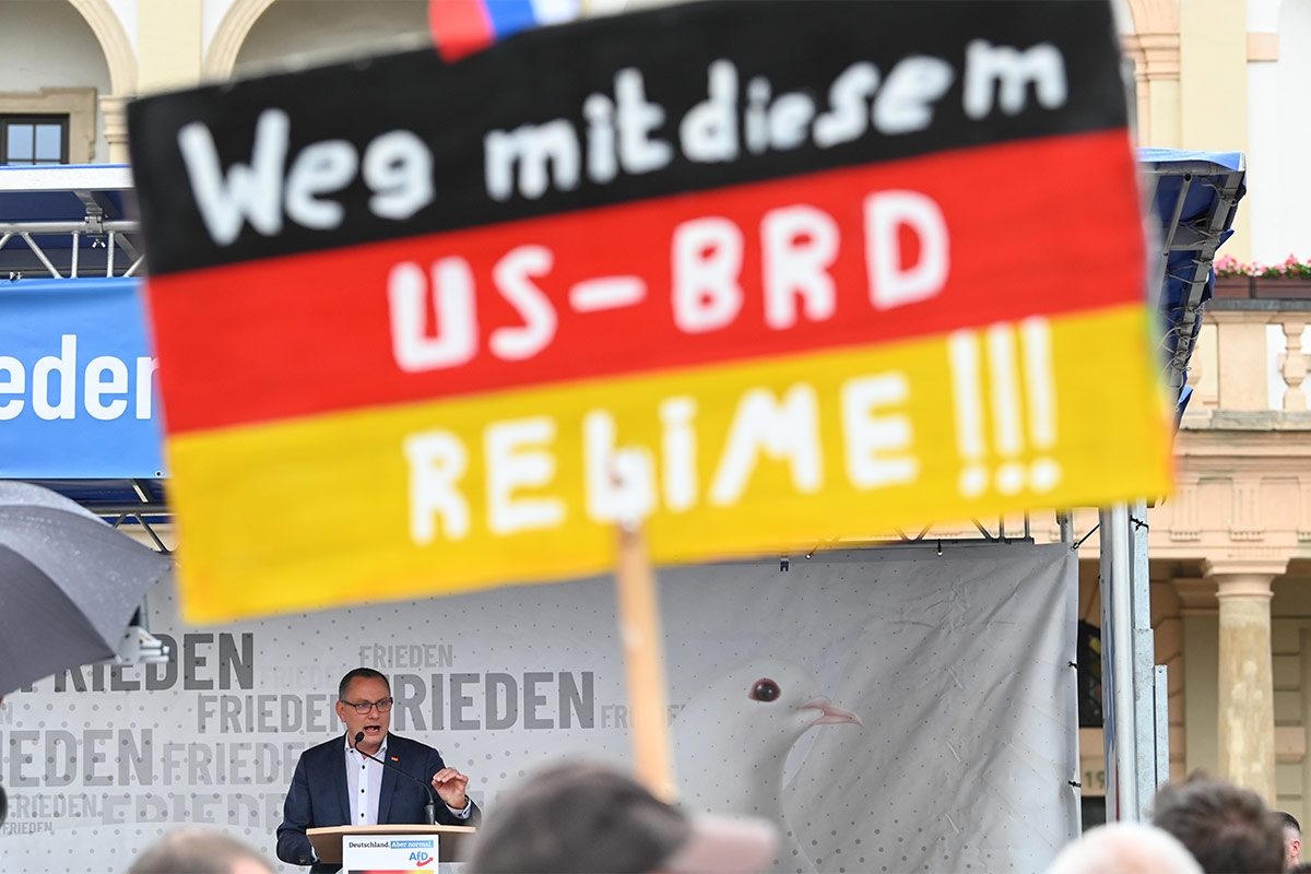 Schild in schwarz-rot-gold mit dem Spruch »Weg mit diesem US-BRD-Regime«, dahinter auf einer Bühne Chrupalla bei einer Rede