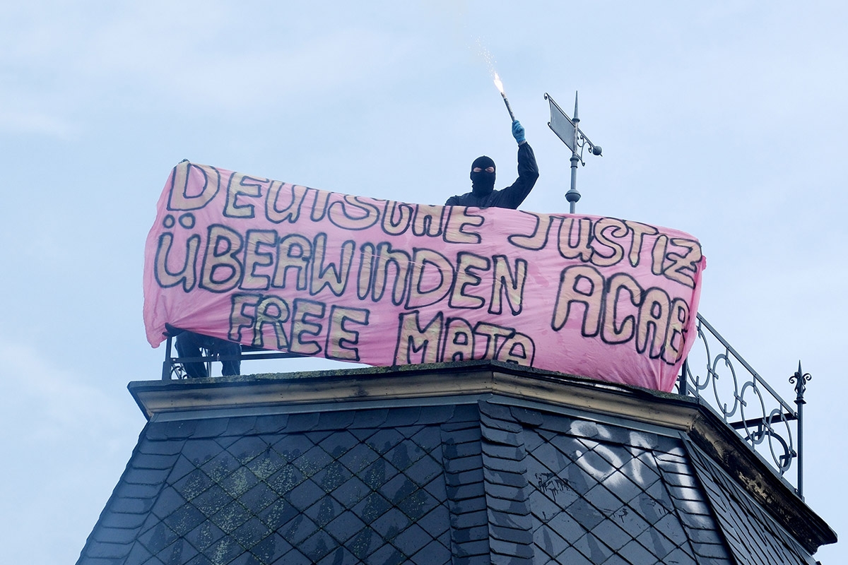 Vermummte Person auf Hausdach, daneben Transpi mit der Aufschrift: "Deutsche Justiz überwinden - ACAB - Free Maja"