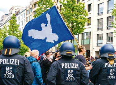 Hinter einer Reihe Polizisten stehen Demonstrant:innen, einer von ihnen trägt eine blaue Fahne mit Friedenstaube
