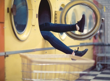 Beine in Jeans mit schwarzen Turnschuhen ragen aus einer großen gelben Waschmaschine in einem Waschsalon