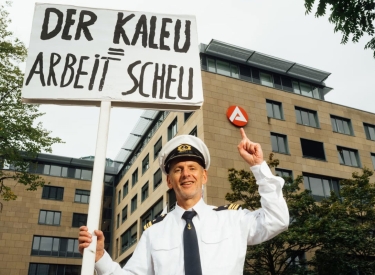 Der Kaleu mit einem Schild mit der Aufschrift »Der Kaleu = Arbeitsscheu«