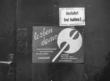 Schwarze Wand mit einem Plakat, auf dem eine Doppelaxt zu sehen ist, daneben der Schriftzug »lesbendemo«, darunter »Gegen Deutschnationalismus und Heterosexismus, für einen starken Lesbenkampf. Lesbendemo, Am 3.11.1990, um 11.00, Treffpunkt: Oranienplatz, Ende: Hermannplatz«